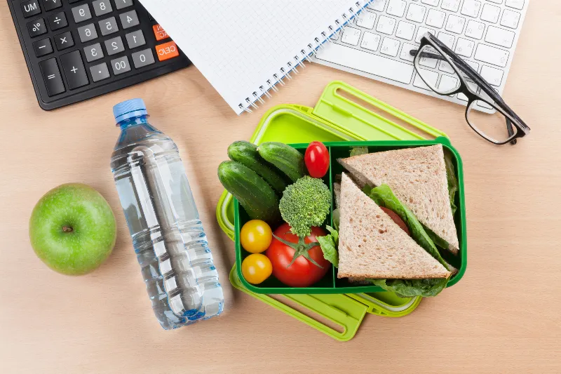 Comida saludable en tupper sobre escritorio con portátil, agua y cuaderno